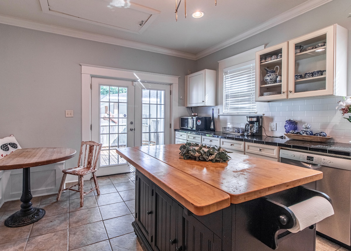 The kitchen features a spacious island with a wooden countertop and dark cabinetry. A round table with a single wooden chair is positioned nearby. Light streams through glass-paned doors leading to an outdoor area, while ample counter space is available for meal preparation.