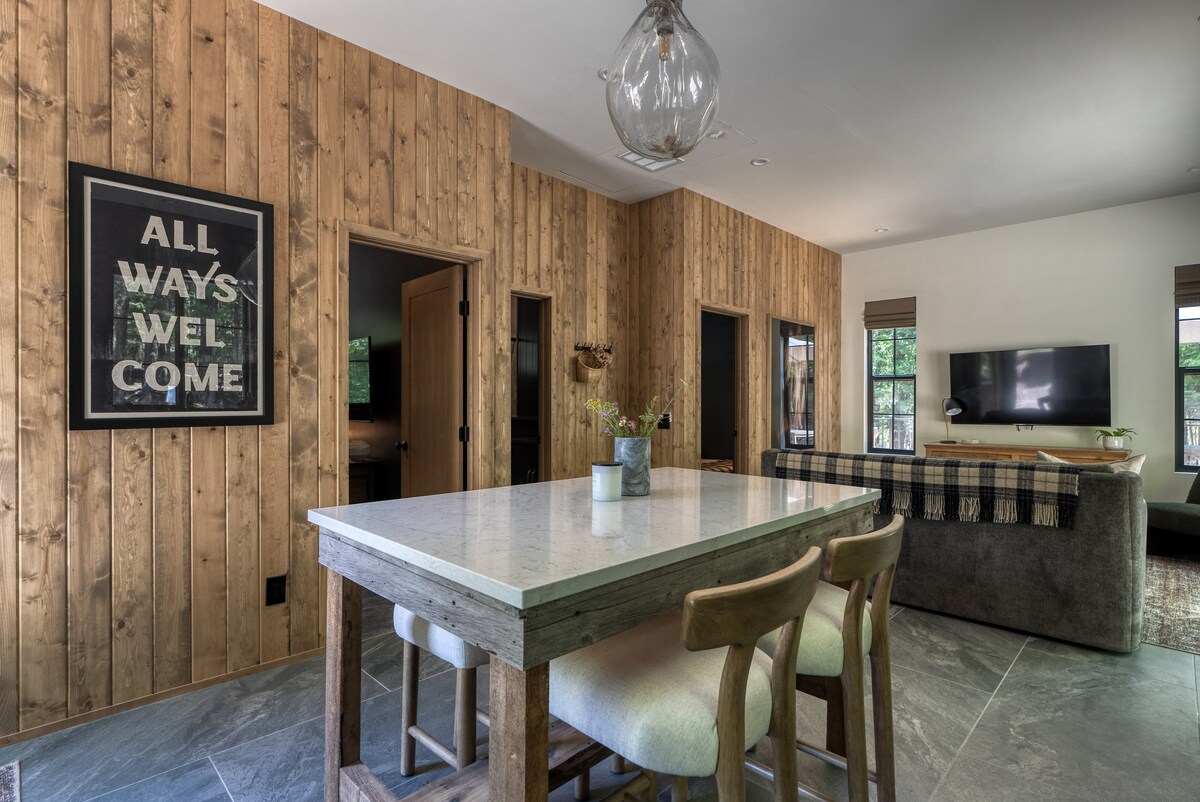 A spacious dining area features a light-colored table with seating for four, surrounded by warm wood-paneled walls. A modern light fixture hangs above, while a framed sign reading 'ALL WAYS WELCOME' is displayed on the wall. A comfortable seating area with a TV is visible in the background.