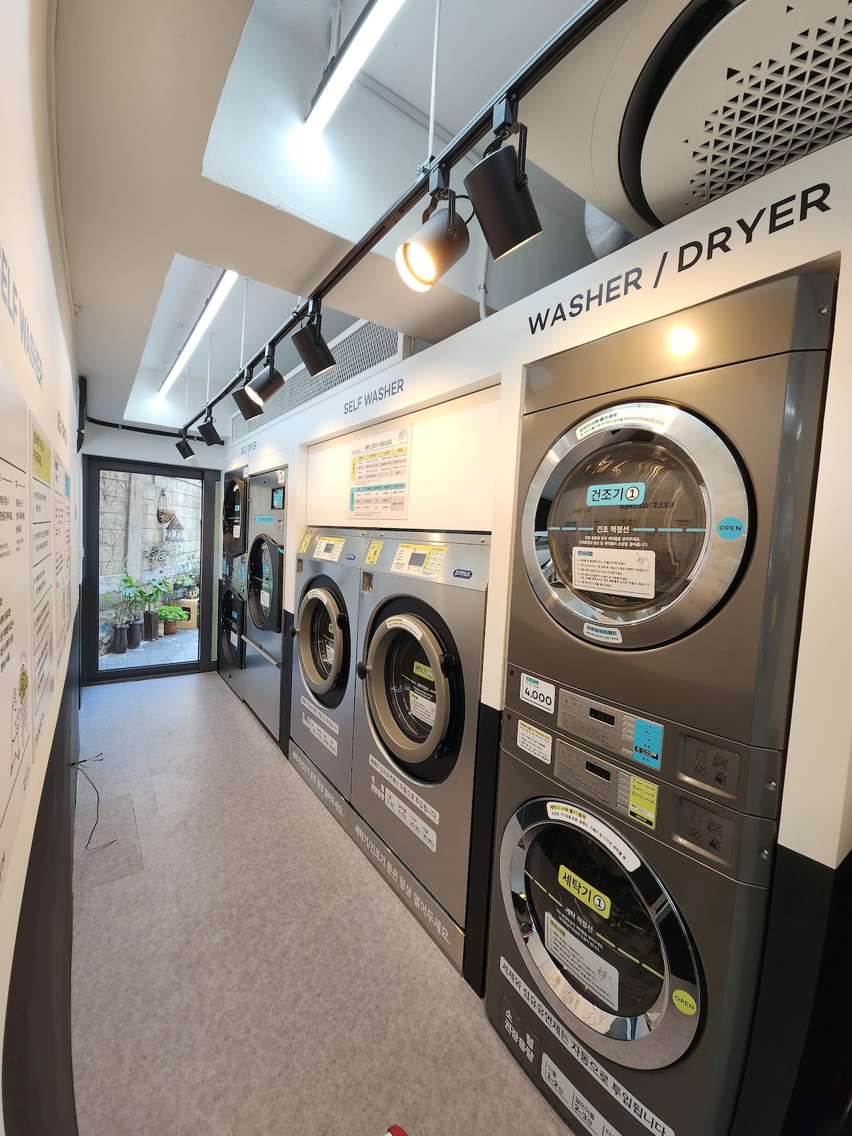 The laundry area features modern self-service washers and dryers lined along one side. A well-organized space is illuminated by ceiling lights, and clear signage describes usage instructions. Potted plants can be seen in the background, adding a touch of greenery to the environment.