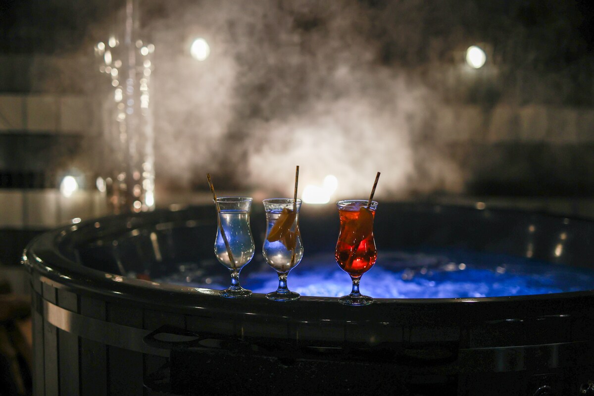 Three colorful cocktails are displayed in elegant glasses by the edge of a bubbling, warm bath. Soft steam rises around, creating a cozy atmosphere. Dim lighting and glowing elements enhance the serene setting, perfect for relaxation.