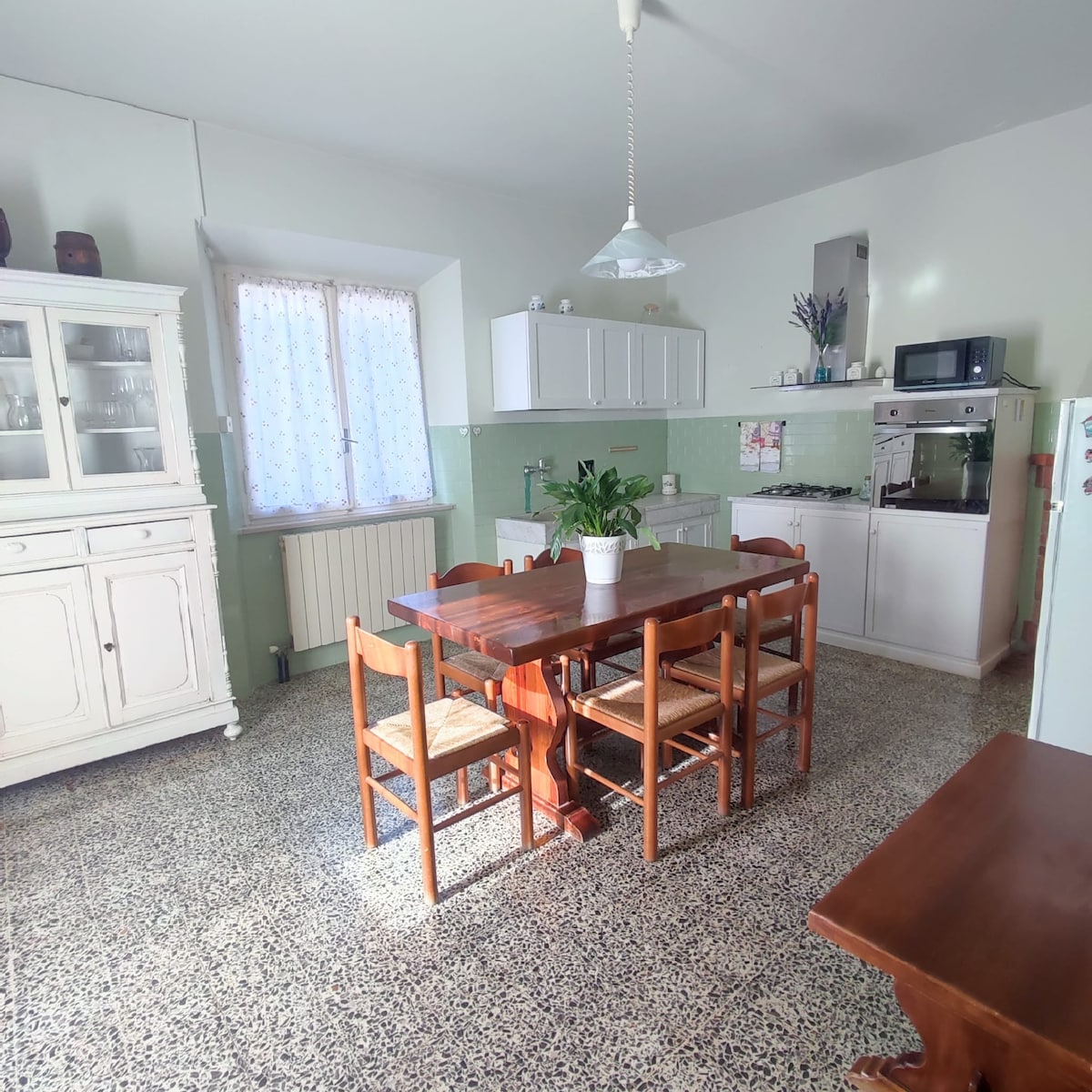 A spacious kitchen is presented, featuring a wooden dining table surrounded by four chairs. A large window allows natural light to fill the room. Appliances, including a microwave and stove, are visible alongside ample counter and storage space.