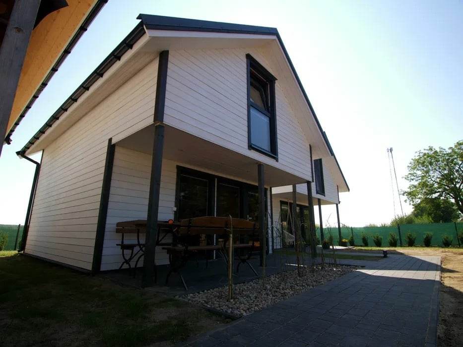 A modern two-story house is depicted, featuring white horizontal siding and large windows. A patio area with outdoor tables is visible, surrounded by landscaped grass. The structure is complemented by a clear blue sky and greenery in the background.