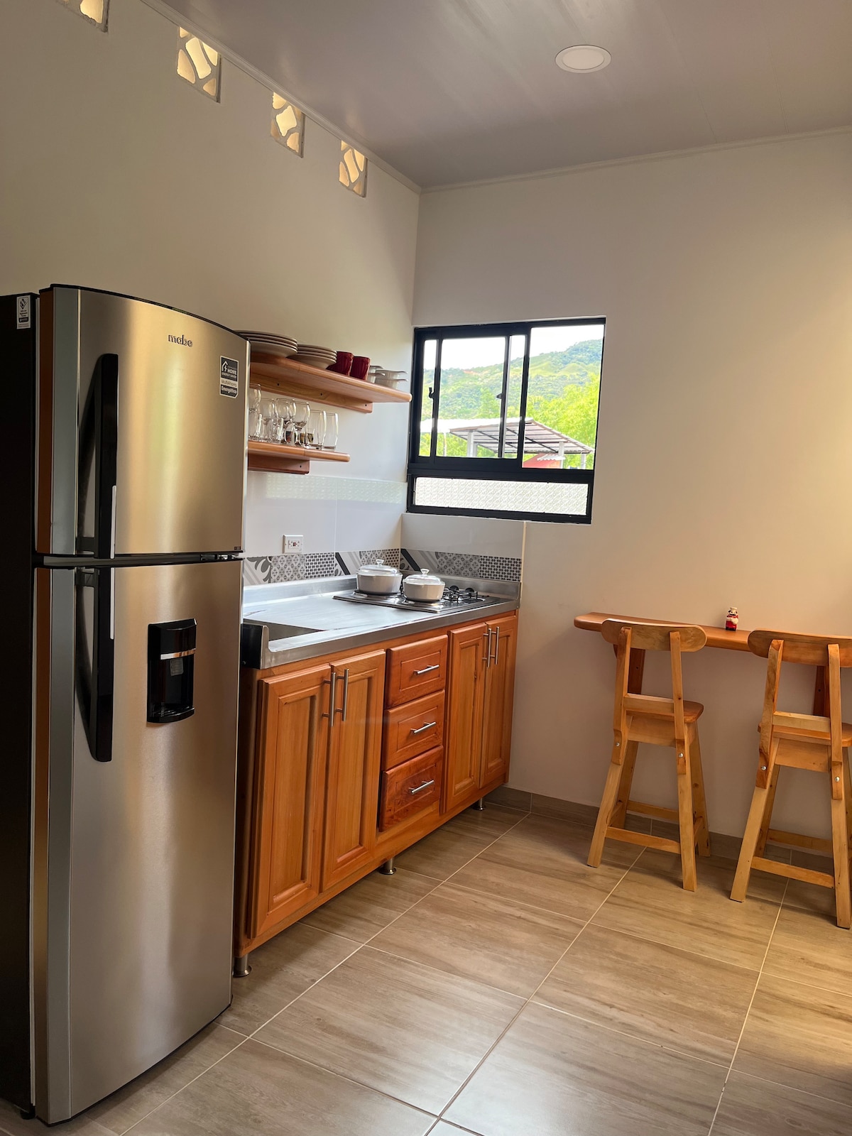 The kitchen showcases sleek stainless steel appliances, including a refrigerator and a gas stove. Wooden cabinets provide storage space, while a small table with two stools offers a cozy dining area. Natural light enters through a window overlooking the surroundings.