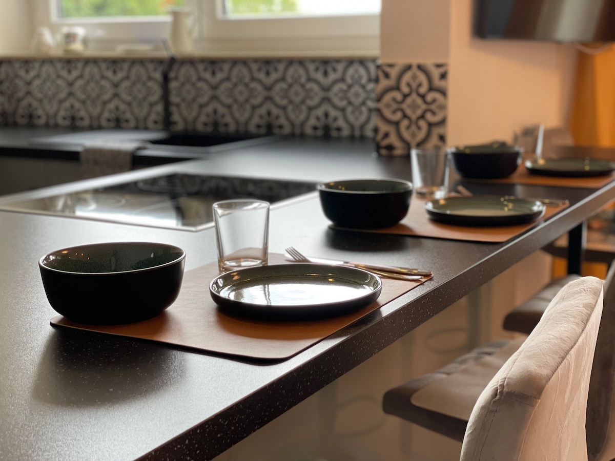 A modern kitchen space features a sleek countertop set for four, including dark green bowls, plates, glasses, and cutlery. Elegant placemats add a refined touch, while the patterned backsplash enhances the contemporary design of the area.
