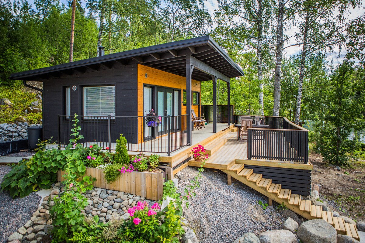 A modern cottage nestled among trees showcases a wooden deck with seating areas, surrounded by a landscaped garden featuring colorful flowers. The structure features large windows allowing natural light, while the approach is enhanced by wooden steps leading to the entrance.