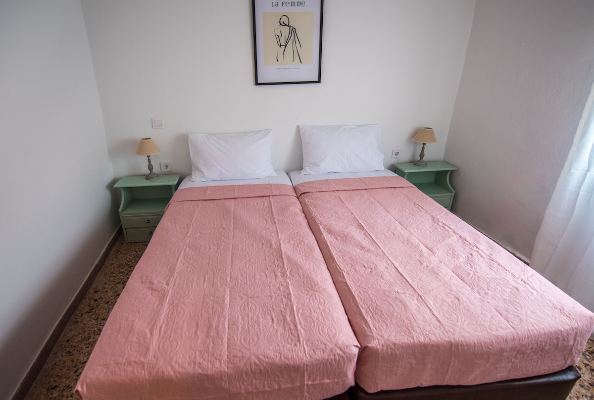 A well-lit bedroom features two spacious beds with pink bedspreads. Each bed is accompanied by a small green bedside table with a lamp. A minimalistic wall art piece is displayed above the beds, complementing the clean, bright walls.