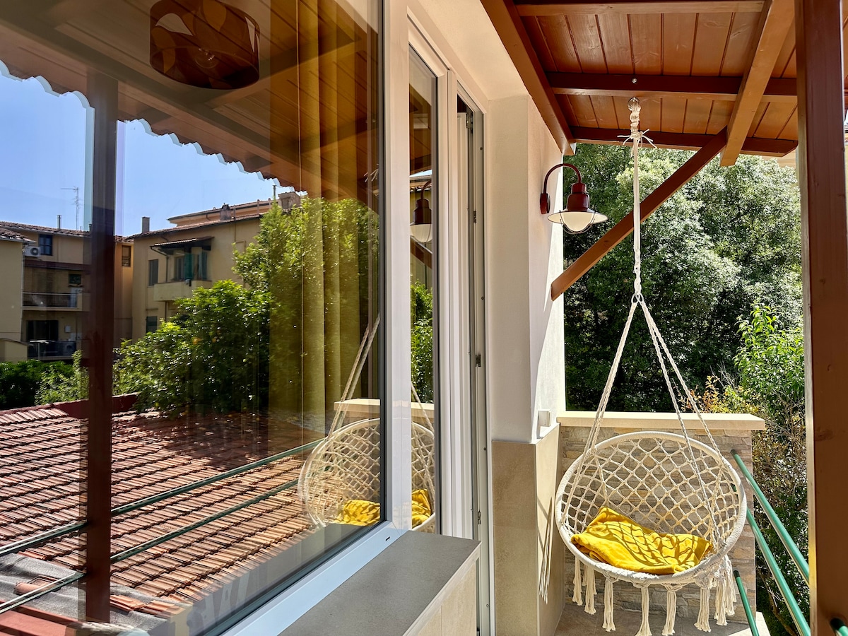 A private balcony is visible, featuring a hanging chair with a yellow cushion. Sunlight reflects off the glass doors, illuminating the space. Greenery can be seen in the background, providing a peaceful view. The balcony's wooden overhang adds a touch of warmth.