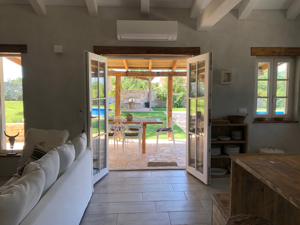Double doors open to a spacious outdoor area, revealing a well-maintained garden and pool. Natural light fills the interior, highlighting a comfortable seating arrangement with a neutral, soft sofa and a wooden dining area. Scenic views of greenery can be seen through the doors.