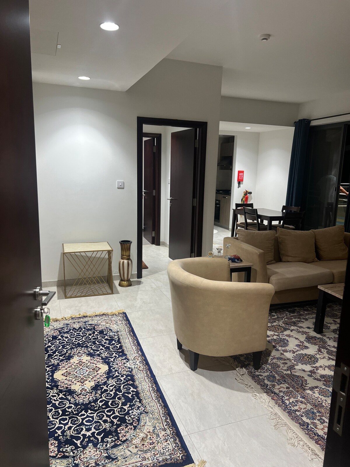 A well-furnished living area features a curved beige sofa and a patterned area rug. A small decorative table is positioned beside the sofa, with a flower arrangement visible in the background. Doorways lead into other rooms, adding depth to the home.