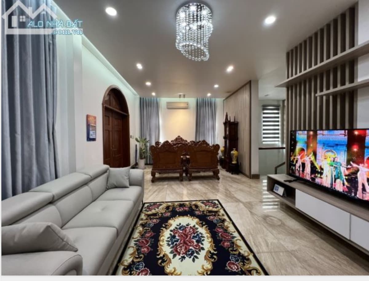 A spacious living area features a light-colored sofa and decorative armchairs. A vibrant area rug with floral designs adds color to the space. A large television is mounted on the wall, complemented by a modern chandelier overhead and stylish curtains framing the windows.