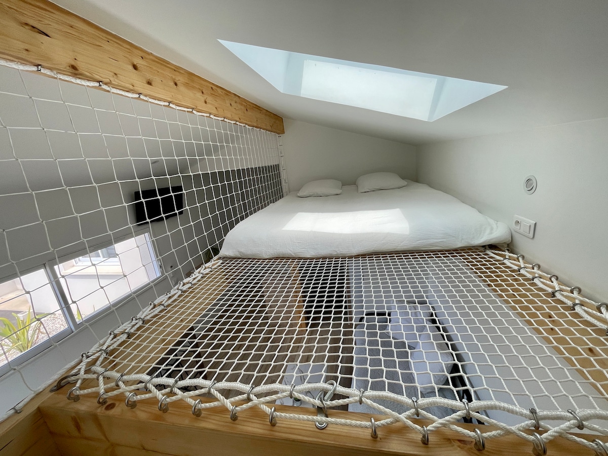 A cozy sleeping area is located on a mezzanine level, featuring a spacious bed with soft bedding and a suspended safety net. A skylight above allows natural light to enter, while windows provide a view of the surrounding space.