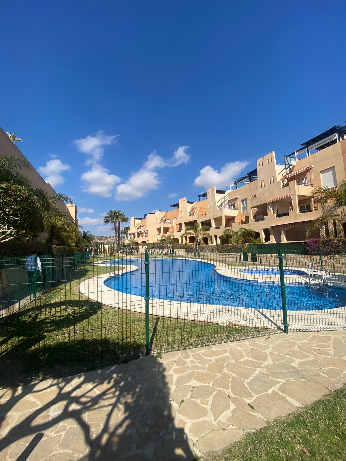 The image captures a serene outdoor area featuring a blue pool, surrounded by landscaped gardens. The pool is enclosed by a fence, with multiple buildings visible in the background, showcasing a harmonious blend of architectural styles against a clear blue sky.