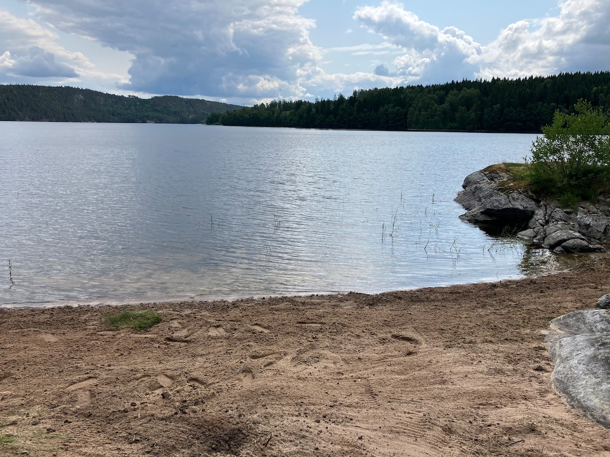 A peaceful lakeside scene is depicted, with smooth, sandy shores leading to calm waters. Dense green trees are visible along the shoreline, framed by a partly cloudy sky. Natural rocky formations border the edge of the lake, creating a serene and private atmosphere.