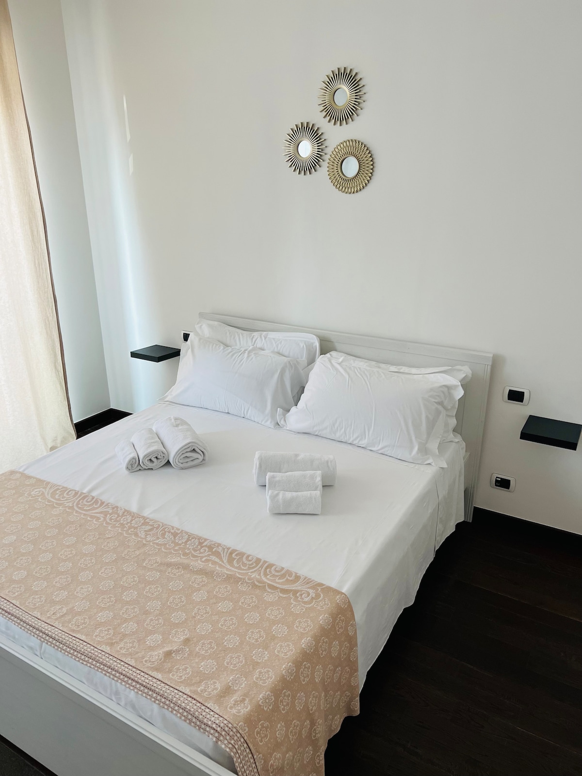 A bright sleeping area features a spacious bed covered with a light blanket. Fresh towels are neatly arranged on the bed. Decorative mirrors are mounted on the wall above the bed, while natural light streams in from a nearby window with sheer curtains.