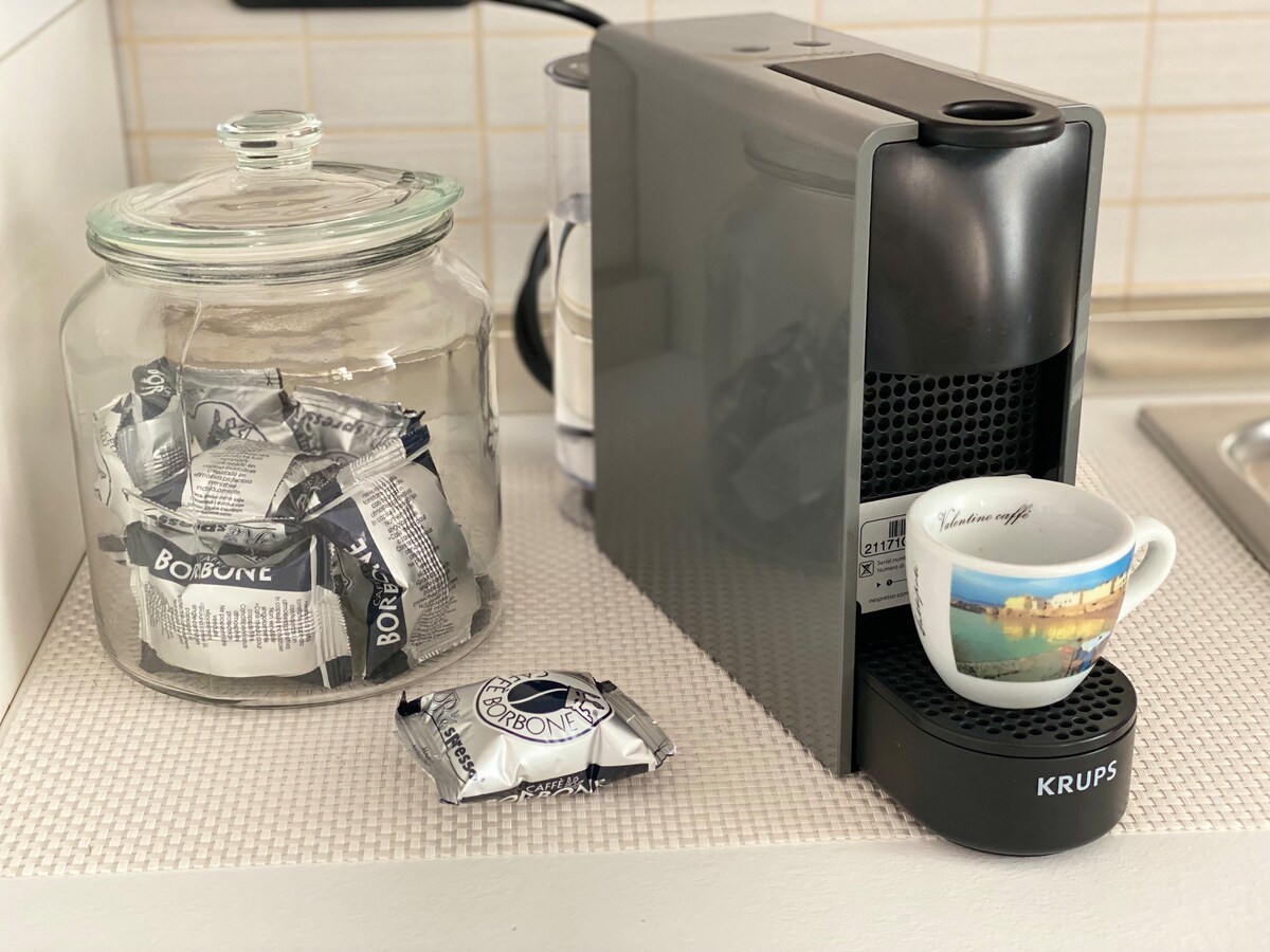 An espresso machine is positioned next to a glass jar filled with coffee capsules. A small coffee cup rests on the machine's tray, ready for use. The countertop appears tidy, and light reflects softly off the machine's surface.