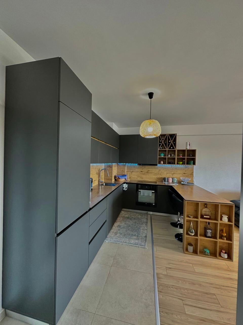 A modern kitchen features sleek black cabinetry and wood accents. A smooth countertop provides workspace and dining options, with bar stools positioned for seating. A variety of bottles and dishes are neatly organized on open shelves, while a window offers natural light to the area.