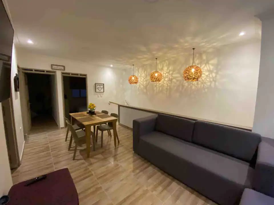 A modern living space is highlighted by a wooden dining table surrounded by four chairs. A spacious gray sofa sits nearby, complemented by intricate pendant lights casting patterned shadows on the walls. Soft lighting contributes to a welcoming atmosphere.