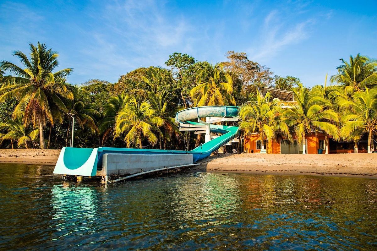 A waterslide extends from a structure and descends into the water, surrounded by palm trees and golden sand. The clear blue sky reflects on the tranquil water, enhancing the natural setting of the eco-lodge by the beach.