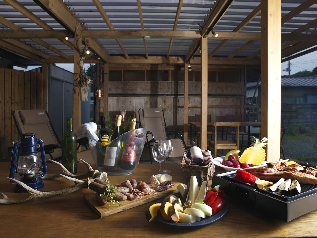 An outdoor dining area is equipped with a wooden table set for a barbecue. Various ingredients, including meats and vegetables, are arranged on a cutting board. Comfortable lounge chairs are positioned nearby, and soft lighting enhances the welcoming ambiance.
