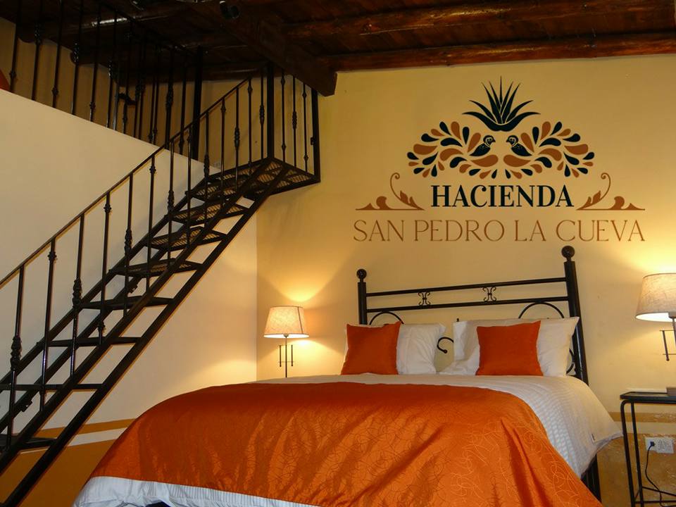 A spacious bedroom features a wrought iron bed adorned with orange and white bedding. A decorative staircase leads to an upper area. Two bedside lamps provide soft illumination, while a decorative wall art element displays the text 'Hacienda San Pedro La Cueva' against a warm yellow backdrop.