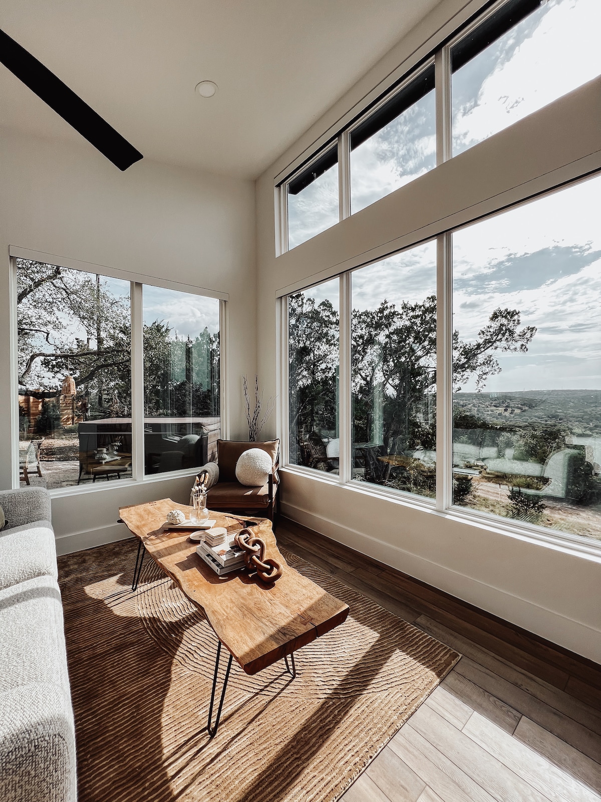 Large windows frame captivating views of the Texas Hill Country, illuminating a bright corner of the living room. A plush sofa complements a rustic wooden coffee table, which is adorned with decorative items. Natural light enhances the warm wood tones of the flooring.