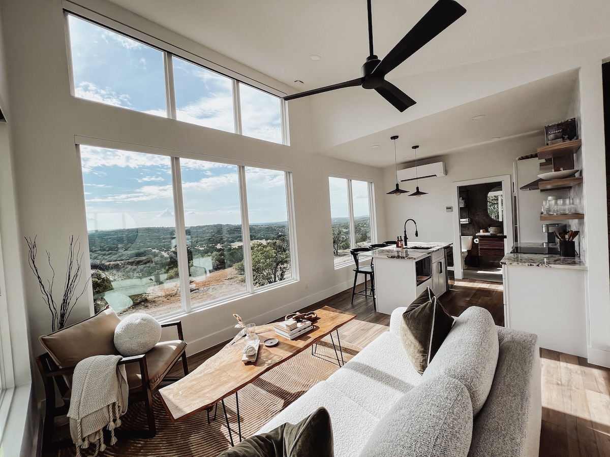 The living room features large windows that provide expansive views of the Texas Hill Country. A spacious sofa and a wooden coffee table are arranged on a textured area rug, creating a comfortable gathering space illuminated by natural light.