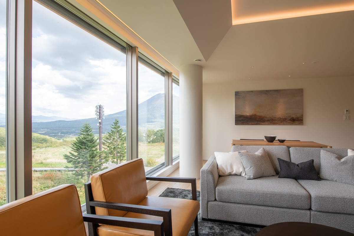 A welcoming living area features large windows showcasing a scenic view of the surrounding landscape, including mountains. Modern furniture includes a cozy gray sofa and two brown armchairs. Soft lighting enhances the space, while a simple piece of wall art completes the setting.