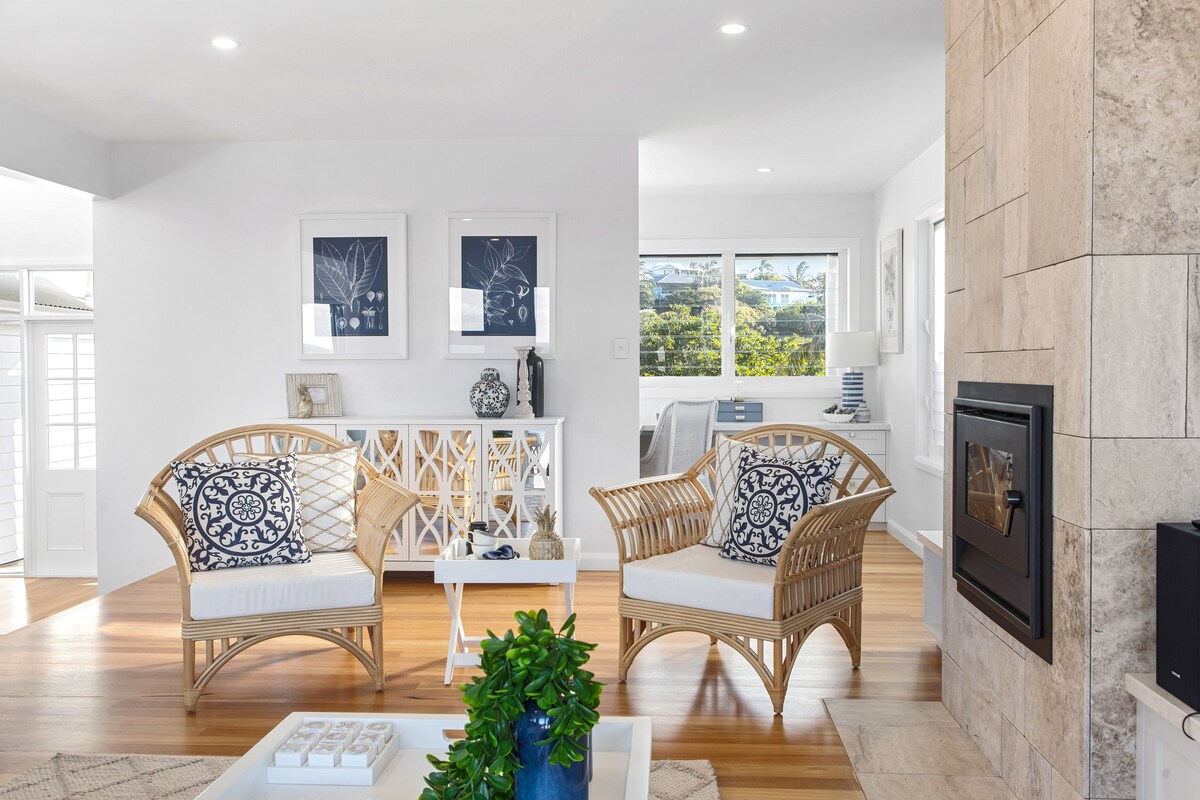 An airy living area features two rattan chairs with decorative cushions, positioned around a low coffee table. A neutral color palette and wooden flooring create a calming atmosphere. Large windows allow natural light to illuminate the space, providing views of the surrounding landscape.