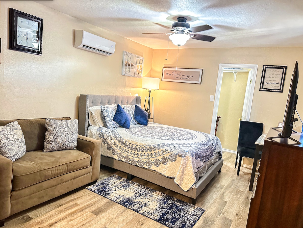 A well-appointed room is presented with a spacious bed adorned with decorative pillows and a patterned blanket. A cozy seating area features a soft sofa, while a desk and TV are positioned nearby. A ceiling fan provides additional comfort in the inviting space.