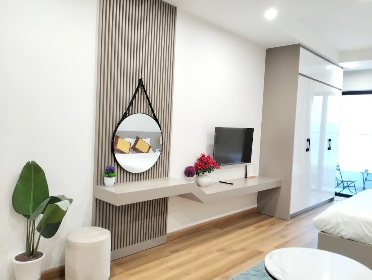 A modern living area features a large round mirror mounted on a textured accent wall. A sleek entertainment unit with a wall-mounted television is complemented by decorative plants and vibrant flowers on the countertop. Light wooden flooring adds warmth to the space.