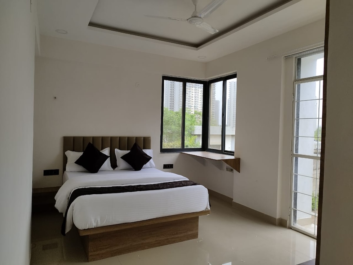 The room features a well-appointed spacious bed with soft linens and two decorative pillows. Large windows provide natural light and an unobstructed view of greenery. A compact work desk is positioned near a window, enhancing the functional layout of the space.