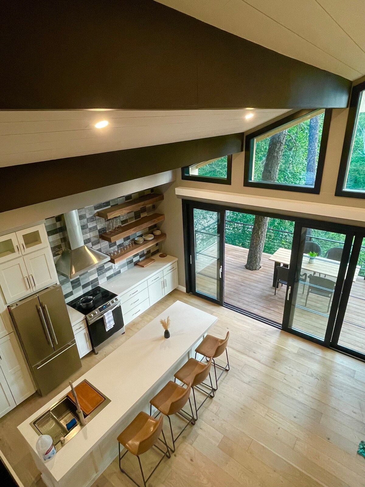 A modern kitchen features sleek white cabinetry and stainless-steel appliances, with a spacious island complemented by four high-backed stools. Large windows provide views of the surrounding trees, allowing natural light to fill the room, while a sliding door leads to a deck area.
