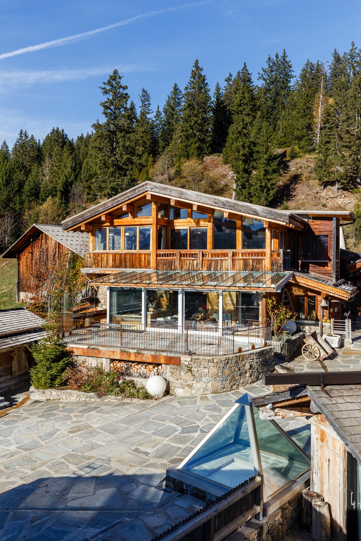 A wooden chalet with expansive glass windows and a spacious stone patio can be seen against a backdrop of lush coniferous trees. The structure displays a rustic design, with warm tones and a natural aesthetic, inviting a connection with the surrounding landscape.