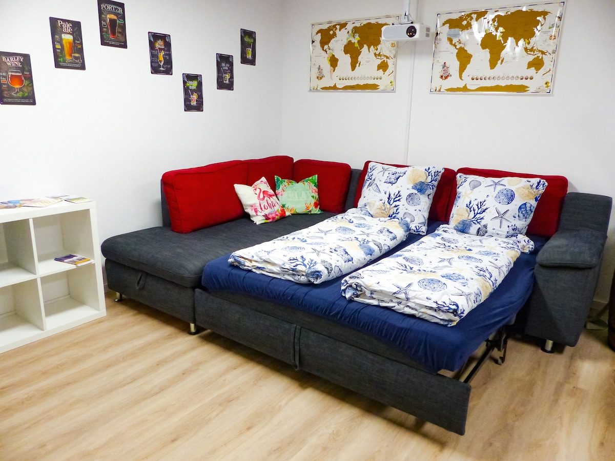 A shared sleeping area features a spacious, convertible couch with two separate beds, each topped with patterned bedding. Colorful cushions add a touch of comfort. A small bookshelf sits nearby, and world map artwork decorates the wall, enhancing the room's ambiance.