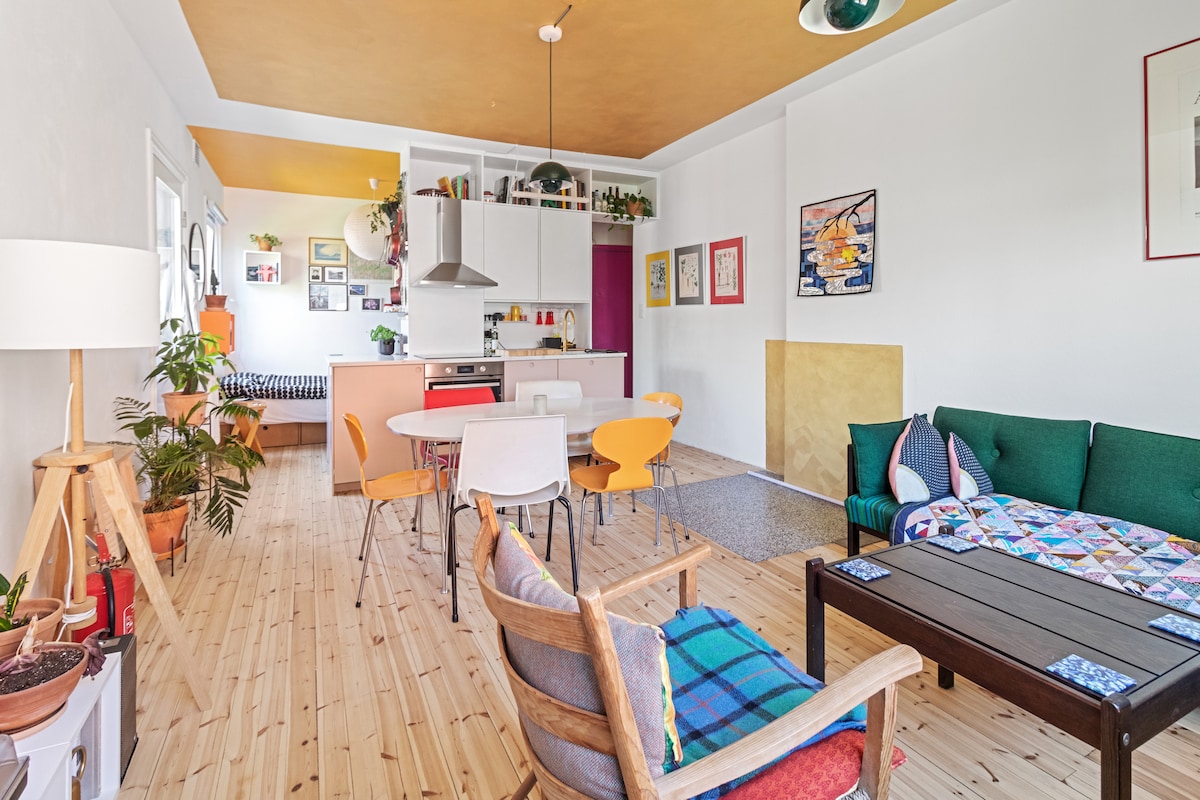 The spacious studio apartment features a well-lit living area with plants and a variety of seating options, including a green couch and wooden chairs. A dining table accommodates several guests, while a bright kitchen area is seen in the background, complemented by colorful decor.
