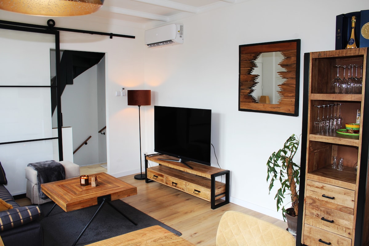 A comfortable living area is presented, featuring a flat-screen television placed on a wooden sideboard. A coffee table made of natural wood sits at the center. A bookshelf holds glassware and decorative items. Soft lighting is provided by a floor lamp, complementing the neutral decor.