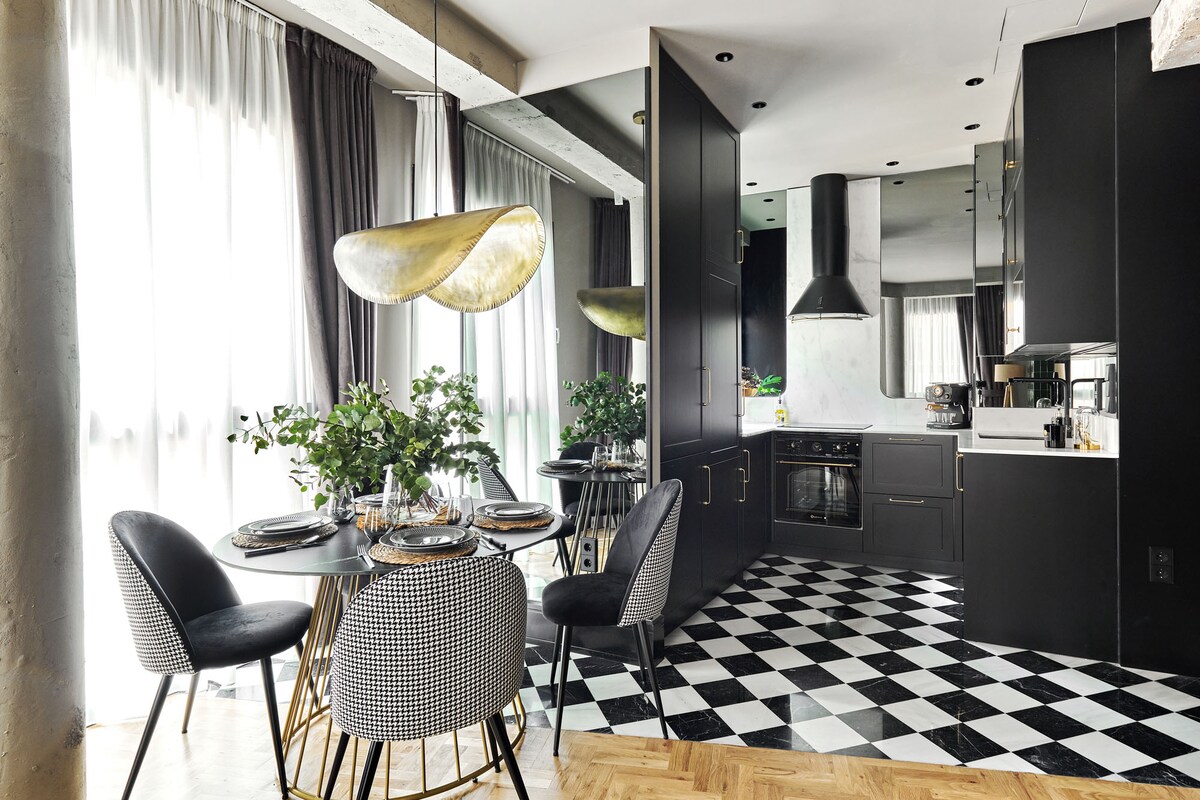 A modern kitchen and dining area is presented, featuring a round dining table surrounded by patterned chairs. Dark cabinetry complements the black and white checkered floor. Natural light is allowed to filter through sheer curtains, enhancing the elegant design of the space.