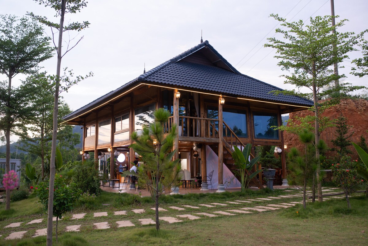 A spacious house featuring a traditional design with a dark tiled roof and large windows. The structure is surrounded by greenery, including tall trees and well-maintained plants, leading to a pathway made of stone. Outdoor seating is visible on the porch.