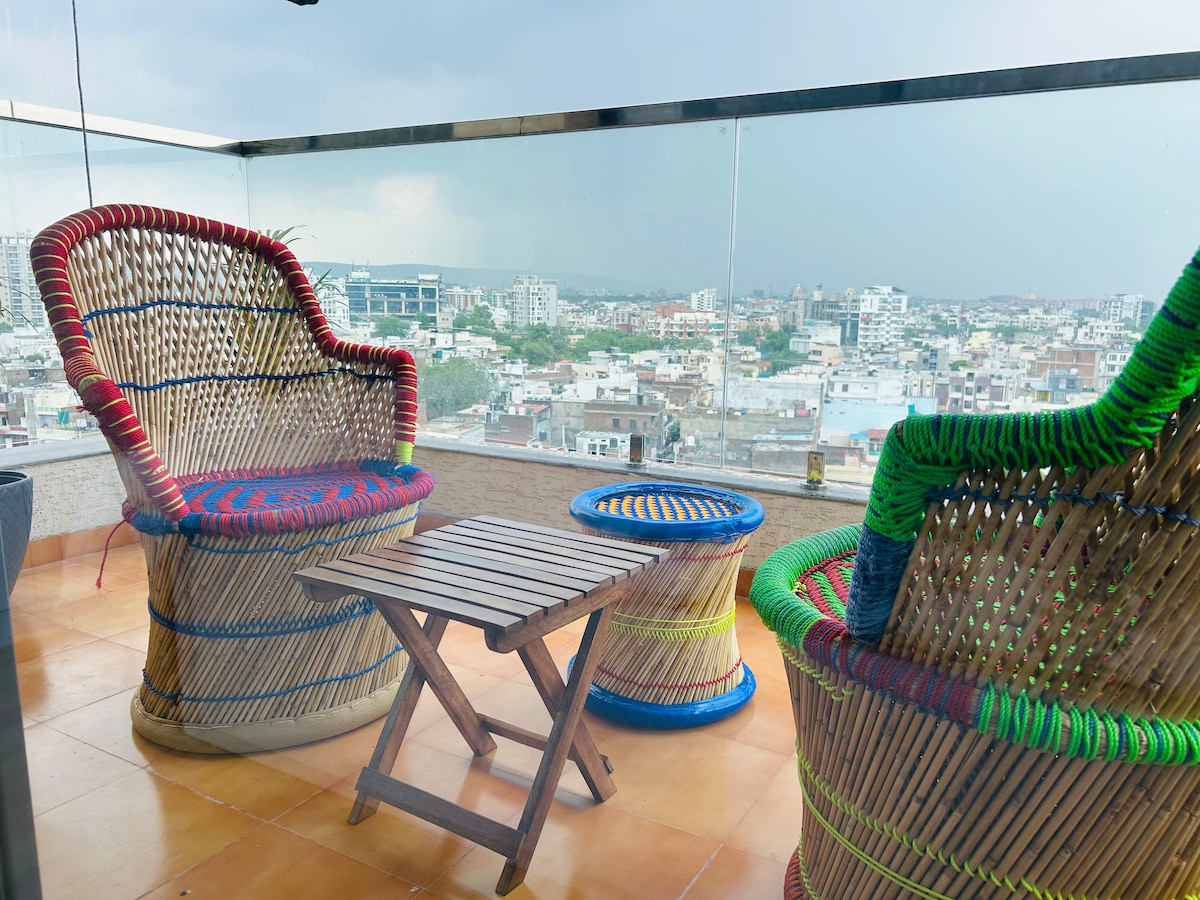 A cozy balcony is furnished with two intricately woven chairs, featuring vibrant colors, and a small wooden table. The space offers unobstructed views of the city skyline, providing an inviting area for relaxation and enjoying the surroundings.