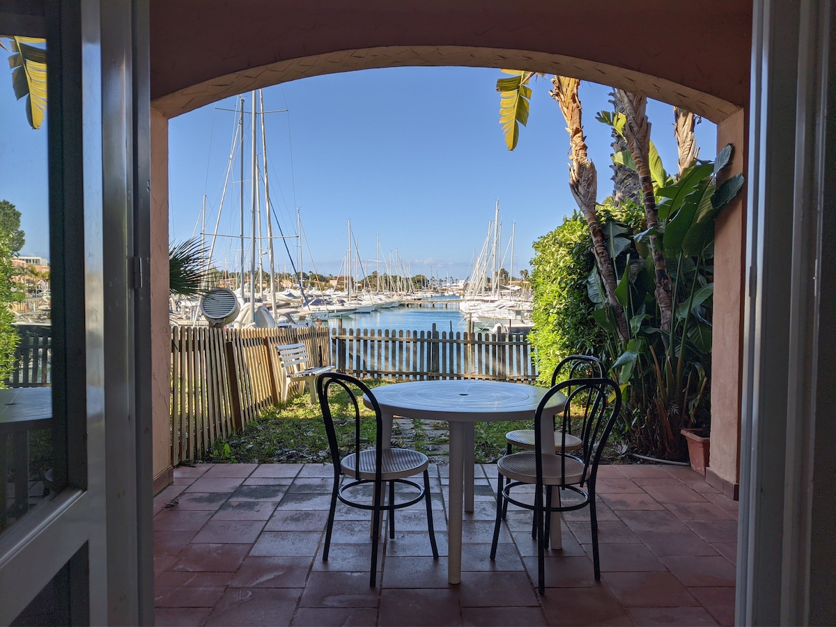 A dining area features a round table accompanied by two chairs, positioned under an archway. Lush greenery surrounds the space, providing a view of the harbor dotted with sailboats. Sunlight filters through, creating a bright and inviting atmosphere.