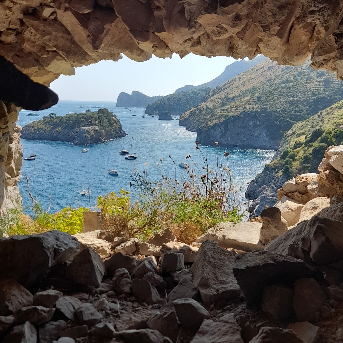 A natural stone opening frames a scenic view of a tranquil bay, where boats are anchored among lush green hills. Sunlight reflects off the water, creating a serene atmosphere enriched by surrounding rocky terrain and coastal vegetation.