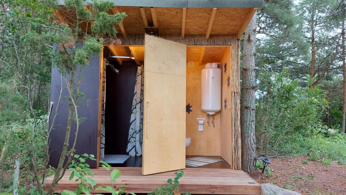 An exterior view of a cozy glamping tent reveals wooden decking and doors that open to a simple interior. The structure features a water heater and a small restroom, surrounded by natural greenery and tall trees, providing a sense of privacy and tranquility.