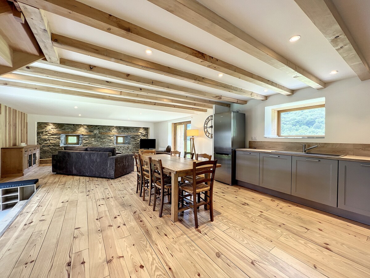 An open-concept living space features wooden beams overhead and a large dining table with eight chairs at the center. Light-colored wooden flooring complements the dark stone accents, while a modern kitchen with sleek cabinetry and stainless-steel appliances is visible in the background. Natural light fills the room through a large window.
