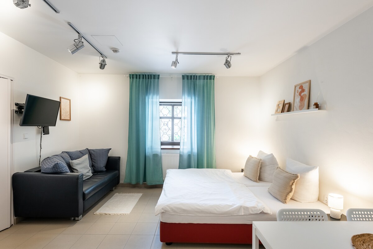 A well-lit room features a spacious bed with light bedding and decorative pillows. A dark sofa sits against one wall, complemented by two accent pillows. Sheer turquoise curtains frame the window, allowing natural light to brighten the space. A small dining table is positioned nearby.