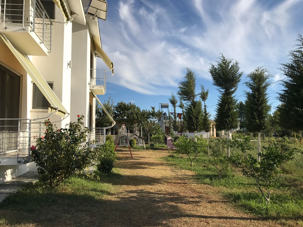 A grassy pathway leads towards a spacious yard, flanked by trees and shrubs. Two residential buildings are visible on one side, while a play area with swings and climbing structures is located in the background, under a sky with scattered clouds.