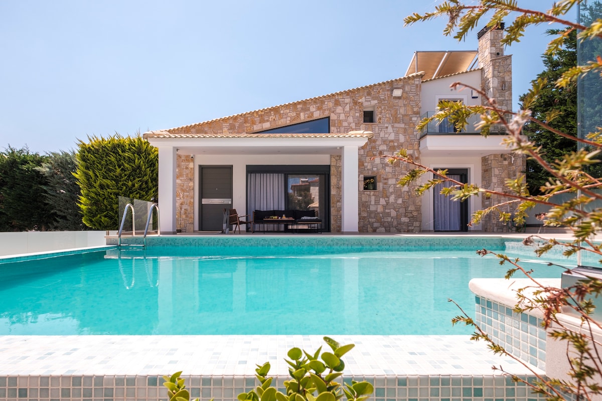 The villa's exterior is highlighted by a large infinity pool, reflecting clear blue water under a bright sky. Stone walls complement the modern architectural design, while shrubs and greenery frame the space, creating a serene and private outdoor environment.