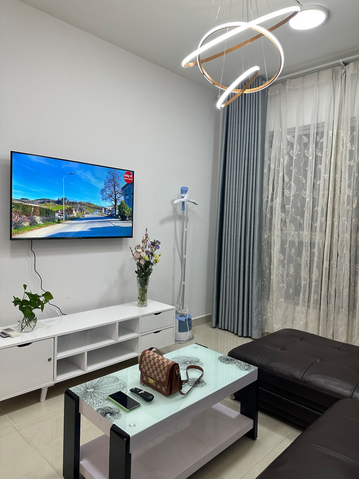 A modern living area features a sleek glass coffee table with a handbag and electronic devices atop it. A comfortable dark sofa invites relaxation, while a television displays a scenic view. Elegant curtains frame the window, and a plant adds a touch of greenery.