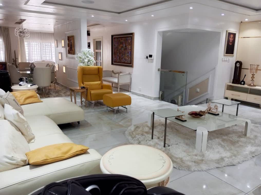 A spacious living room features a light-colored leather sofa adorned with decorative pillows and accented by a plush area rug. A glass coffee table sits centrally, while yellow armchairs add a vibrant touch. Bright natural light fills the space through large windows.