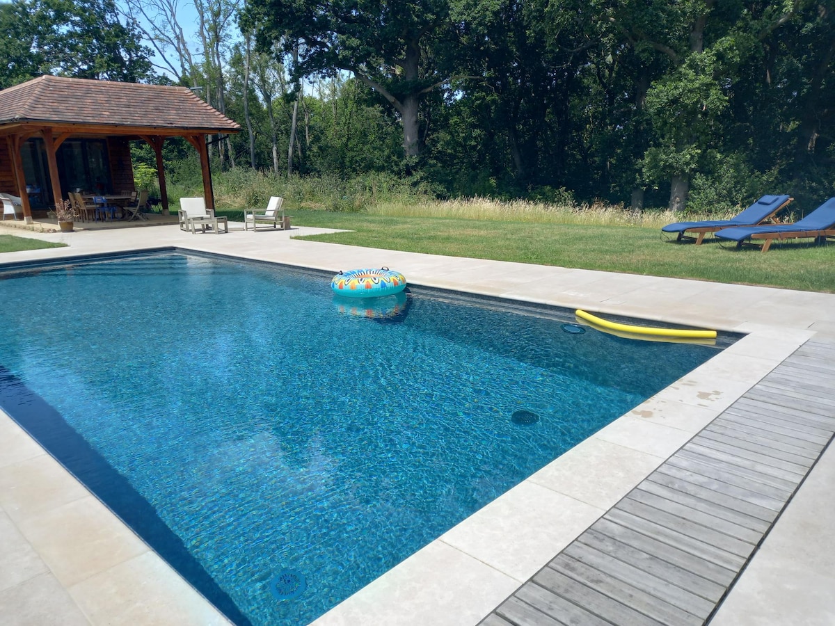 A heated pool is surrounded by a spacious patio, featuring sun loungers and a small covered seating area in the distance. A vibrant inflatable ring floats in the water, with a yellow pool noodle resting on the edge, and lush greenery forms a serene backdrop.