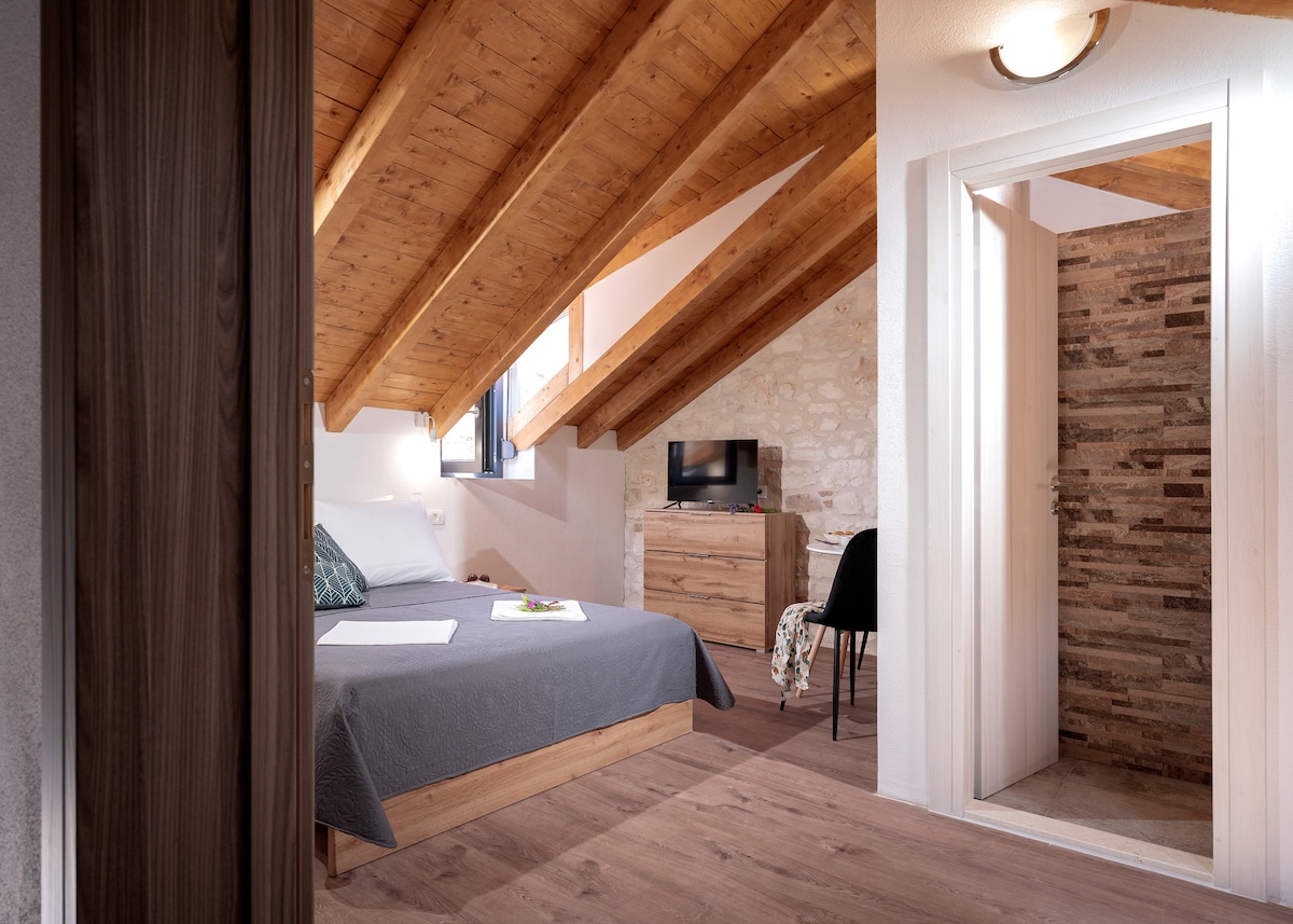 An inviting attic room features a spacious bed against a backdrop of exposed wooden beams. Light streams through the window, illuminating the rustic stone wall. A wooden dresser and a black chair are positioned nearby, while the en-suite bathroom is discreetly accessed through a white door.
